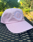 Garment-Washed Logo Hat in pink with Clara & Fritz logo, styled on an outdoor table against a greenery backdrop.