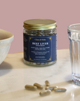 Jar of Beef Liver Supplement capsules on a table next to a bowl and a glass, showcasing nutrient-rich superfood benefits.