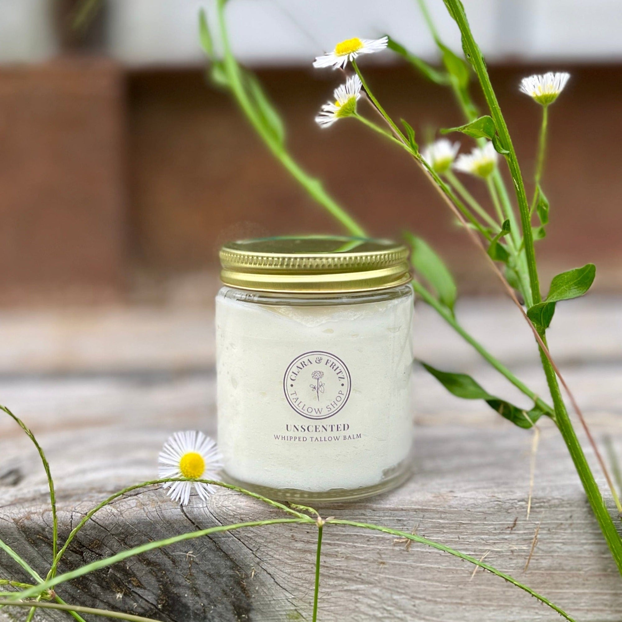 Jar of Clara & Fritz Unscented Whipped Tallow Balm moisturizer, surrounded by flowers, showcasing natural skincare product.