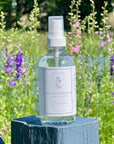 Clear Complexion Refining Facial Mist bottle displayed outdoors with colorful flowers in the background.