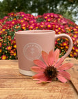 Pink Latte Mug with Clara & Fritz logo on wooden table surrounded by vibrant flowers, perfect for coffee lovers and gifting.