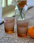 Set of two pink Garden Rose Juice Glasses with orange fruit on a floral tablecloth, showcasing elegant vertical ribbed design.
