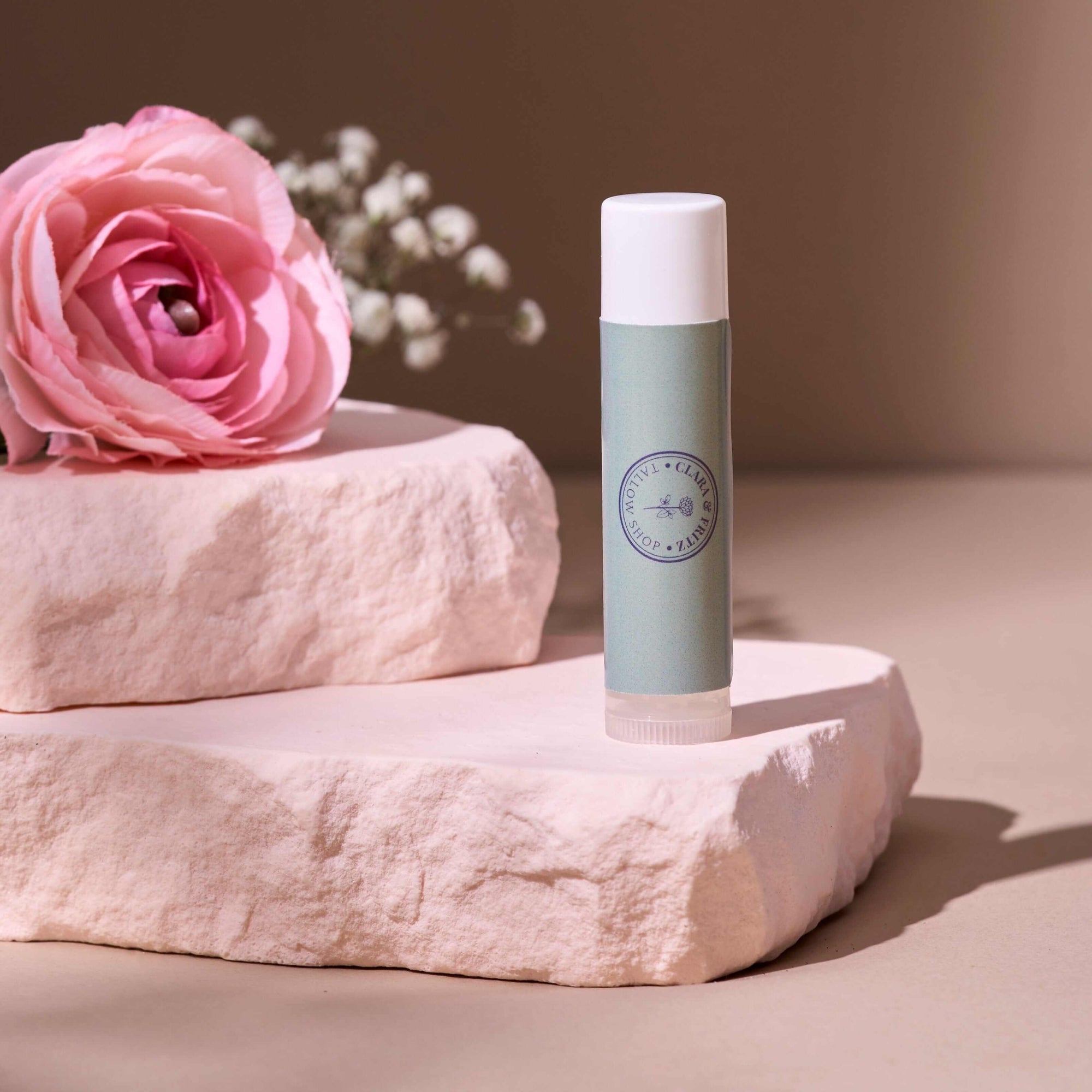 Tallow Lip Balm - Single tube on a textured pink stone with a soft pink flower in the background, offering nutrient-rich hydration. Also included in Lip Balm Set.