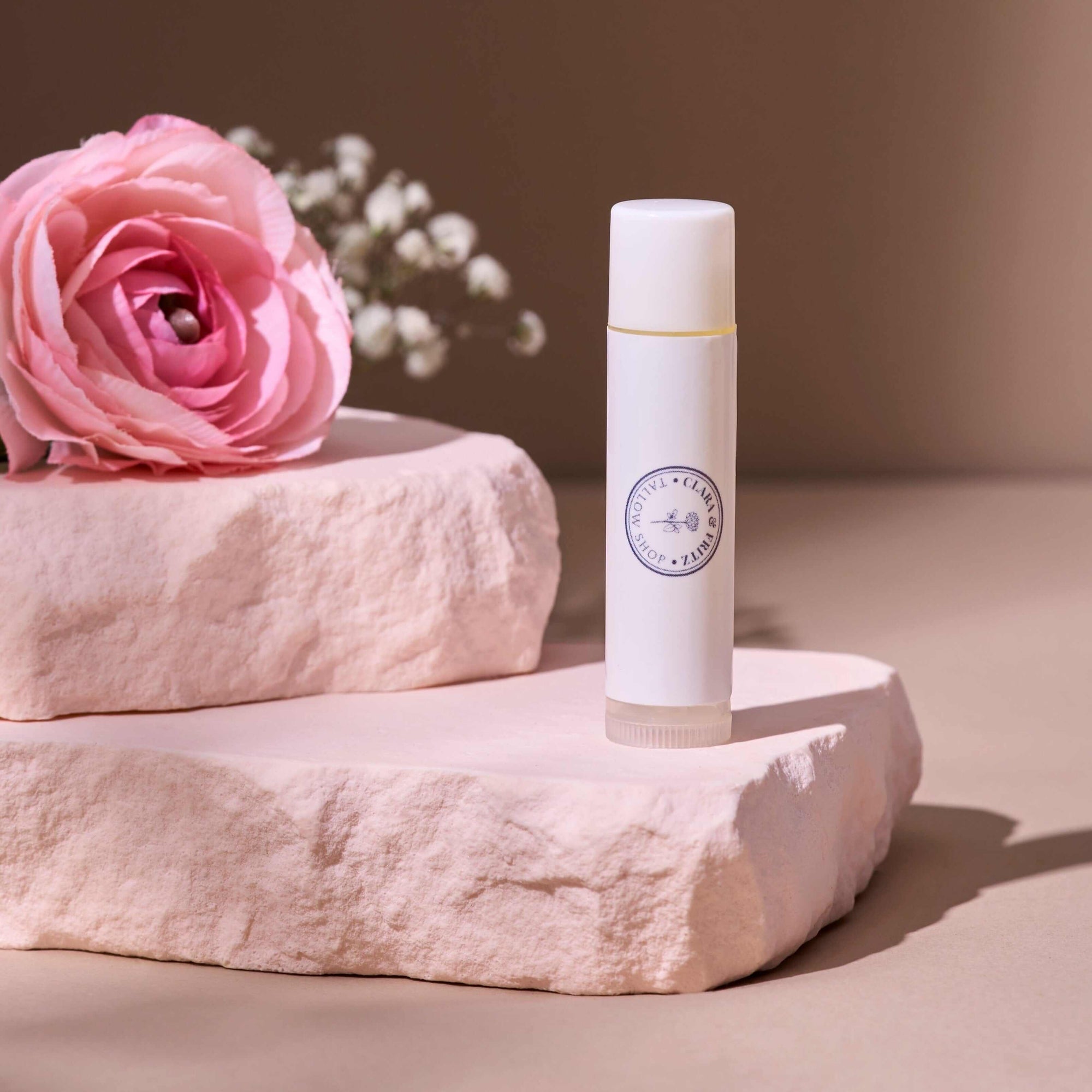 Tallow Lip Balm - Single on a stone platform with pink flowers, offering moisture-rich hydration and a satin finish for all-day wear. Also included in Lip Balm Set.