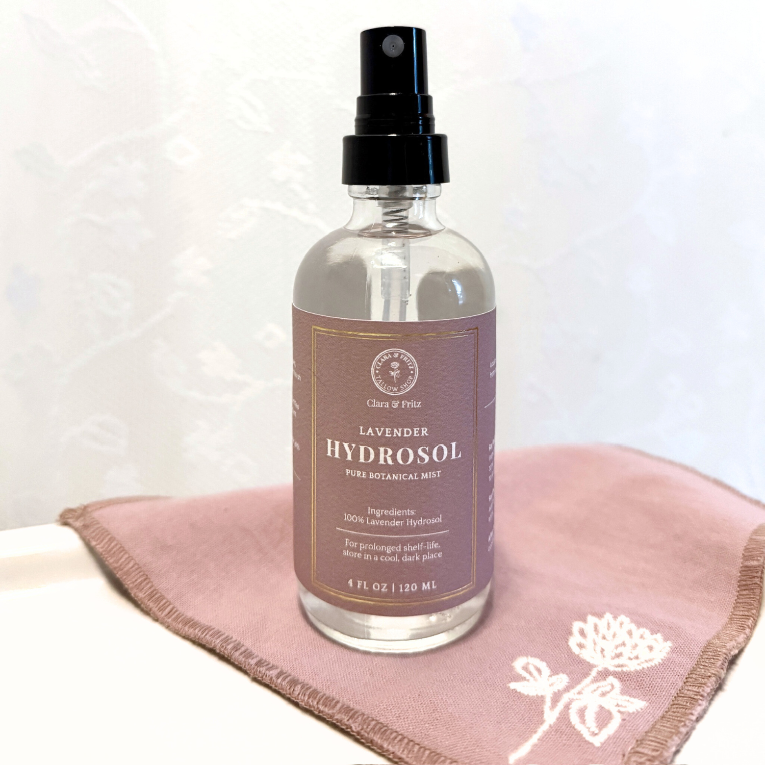 Lavender Hydrosol spray bottle for facial use, displayed on a pink cloth with floral embroidery.