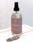 Glass bottle of Lavender Hydrosol for Facial Use with spray nozzle, next to a dried lavender sprig on a white tray