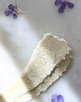 Pink Grapefruit Tallow Sugar Scrub with natural exfoliating texture, surrounded by purple flower petals on a light surface.