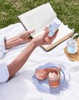 Woman enjoying a picnic with Sun-kissed Trio products, including a skincare bottle and candle, surrounded by fresh fruit and an open book.