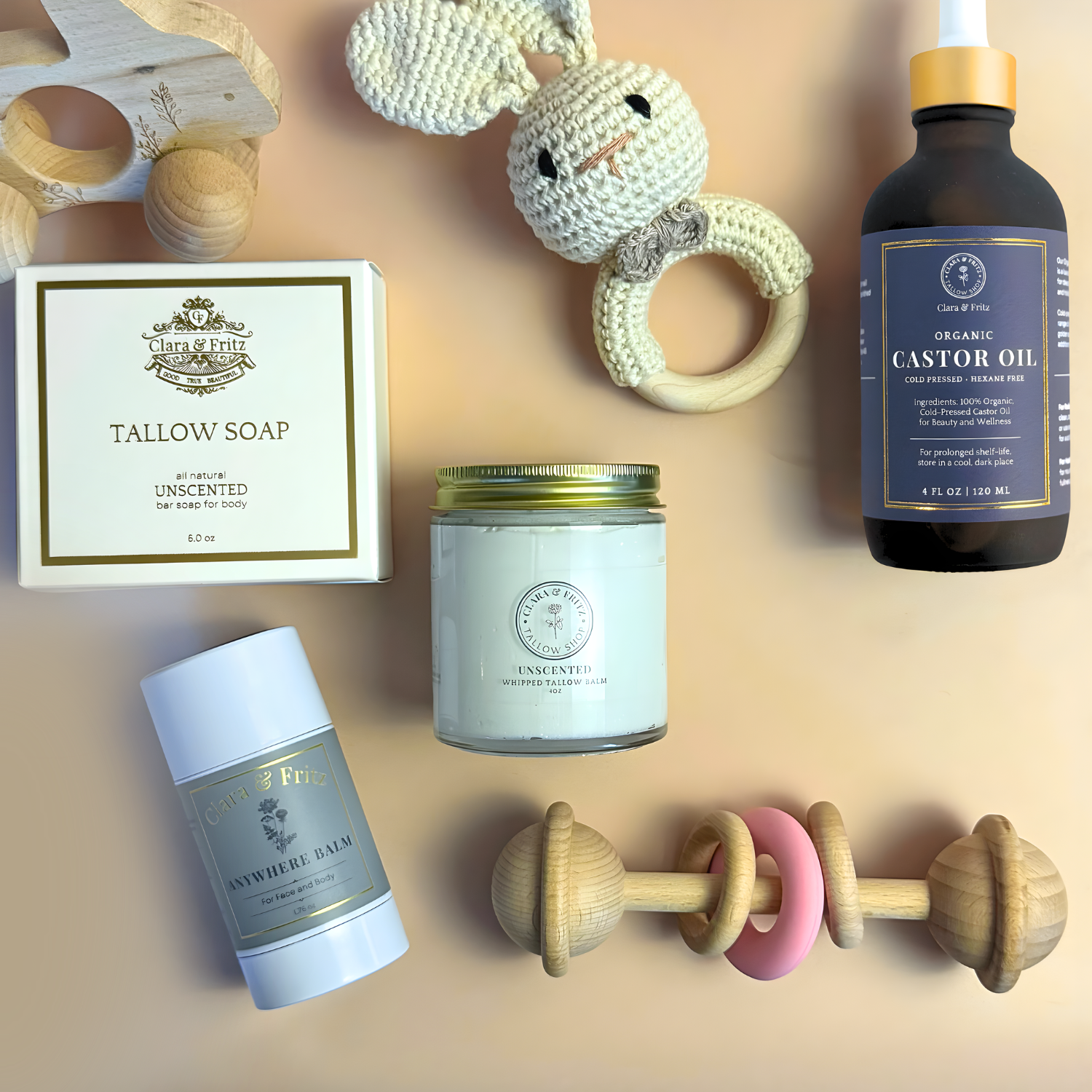Baby Gift Set includes organic castor oil, tallow soap, unscented balm, natural wooden toys, and a knitted bunny rattle on a table.