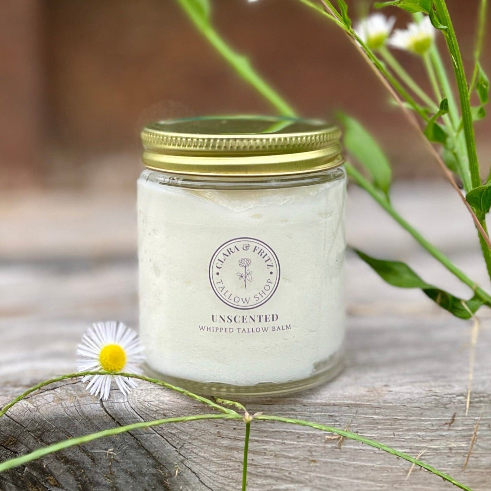 Jar of Clara & Fritz Unscented Whipped Tallow Balm moisturizer, surrounded by flowers, showcasing natural skincare product.