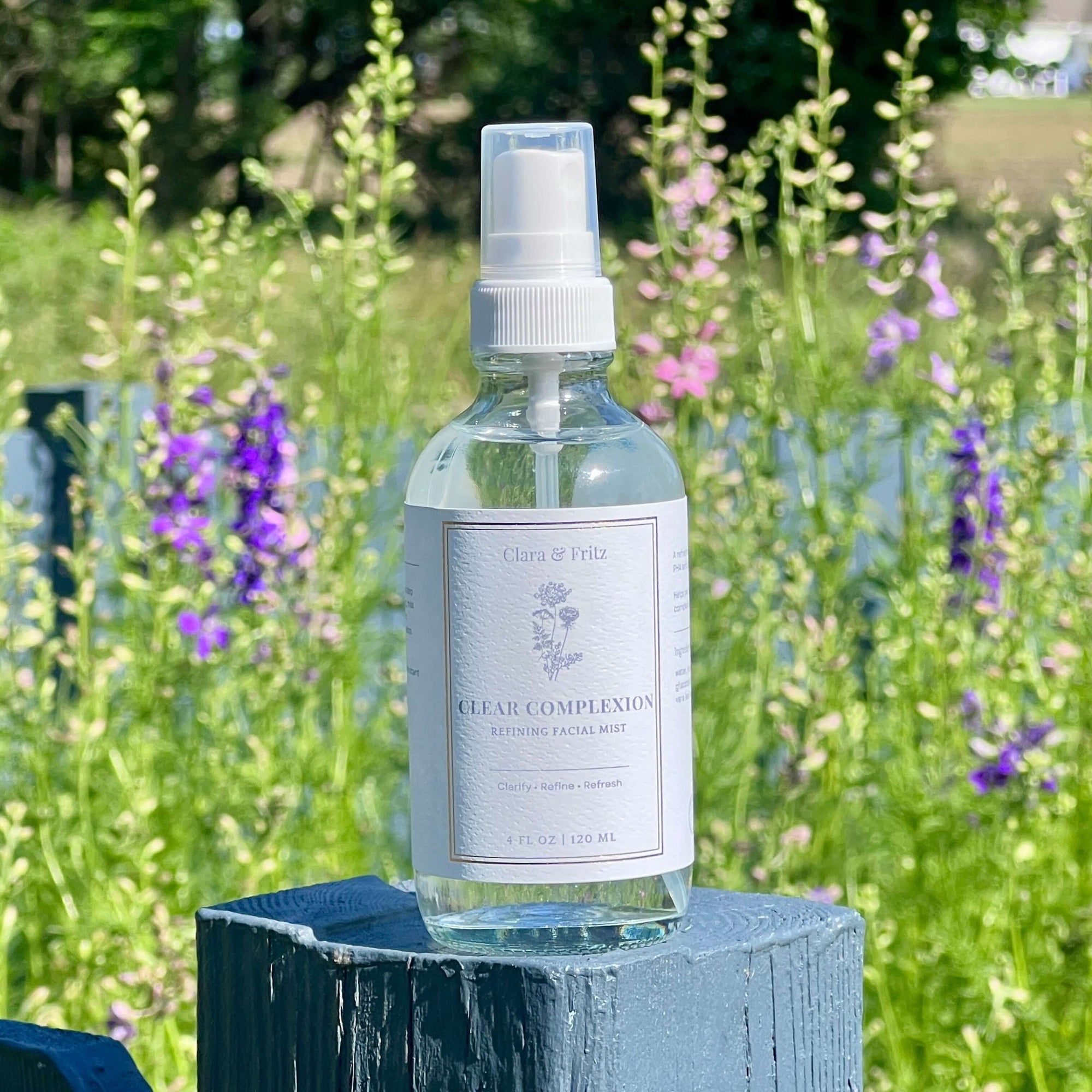 Clear Complexion Refining Facial Mist bottle displayed outdoors with colorful flowers in the background.