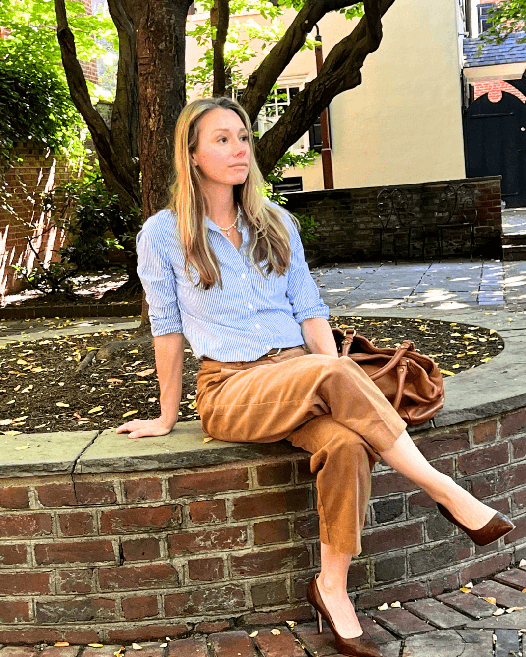 A woman sits on a brick planter in an outdoor park setting, wearing a light blue button-down shirt, brown cropped trousers, and brown heels, with a brown handbag resting beside her.