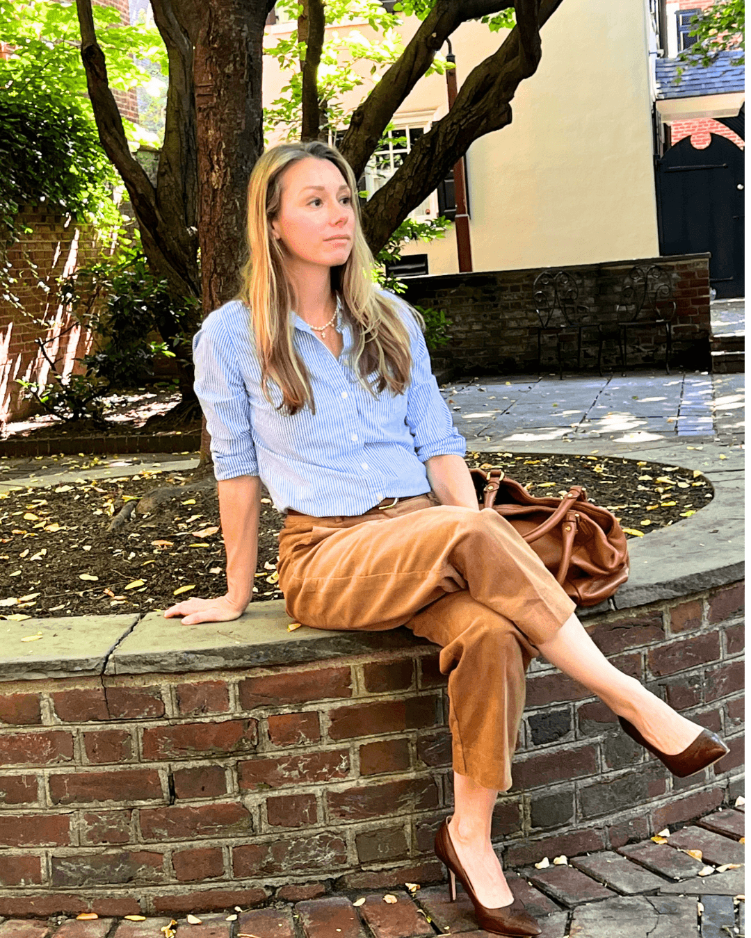 A woman sits on a brick planter in an outdoor park setting, wearing a light blue button-down shirt, brown cropped trousers, and brown heels, with a brown handbag resting beside her.