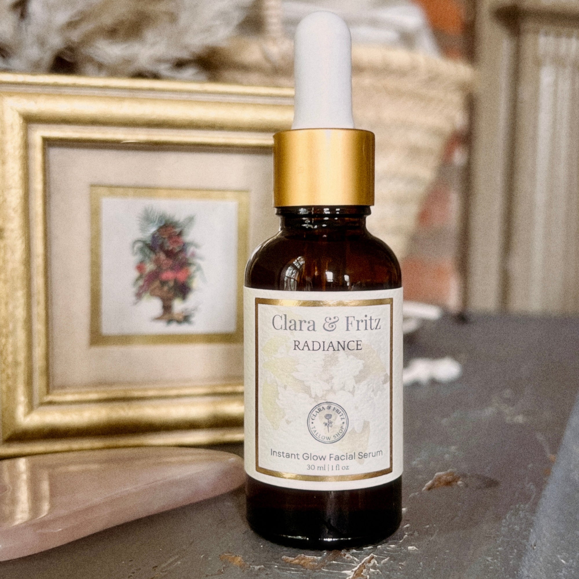 A small amber glass dropper bottle labeled 'Clara & Fritz Radiance Instant Glow Facial Serum' with a white dropper top and gold collar, standing on a stone surface in front of a blurred framed picture.