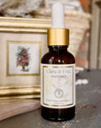 A small amber glass dropper bottle labeled 'Clara & Fritz Radiance Instant Glow Facial Serum' with a white dropper top and gold collar, standing on a stone surface in front of a blurred framed picture.