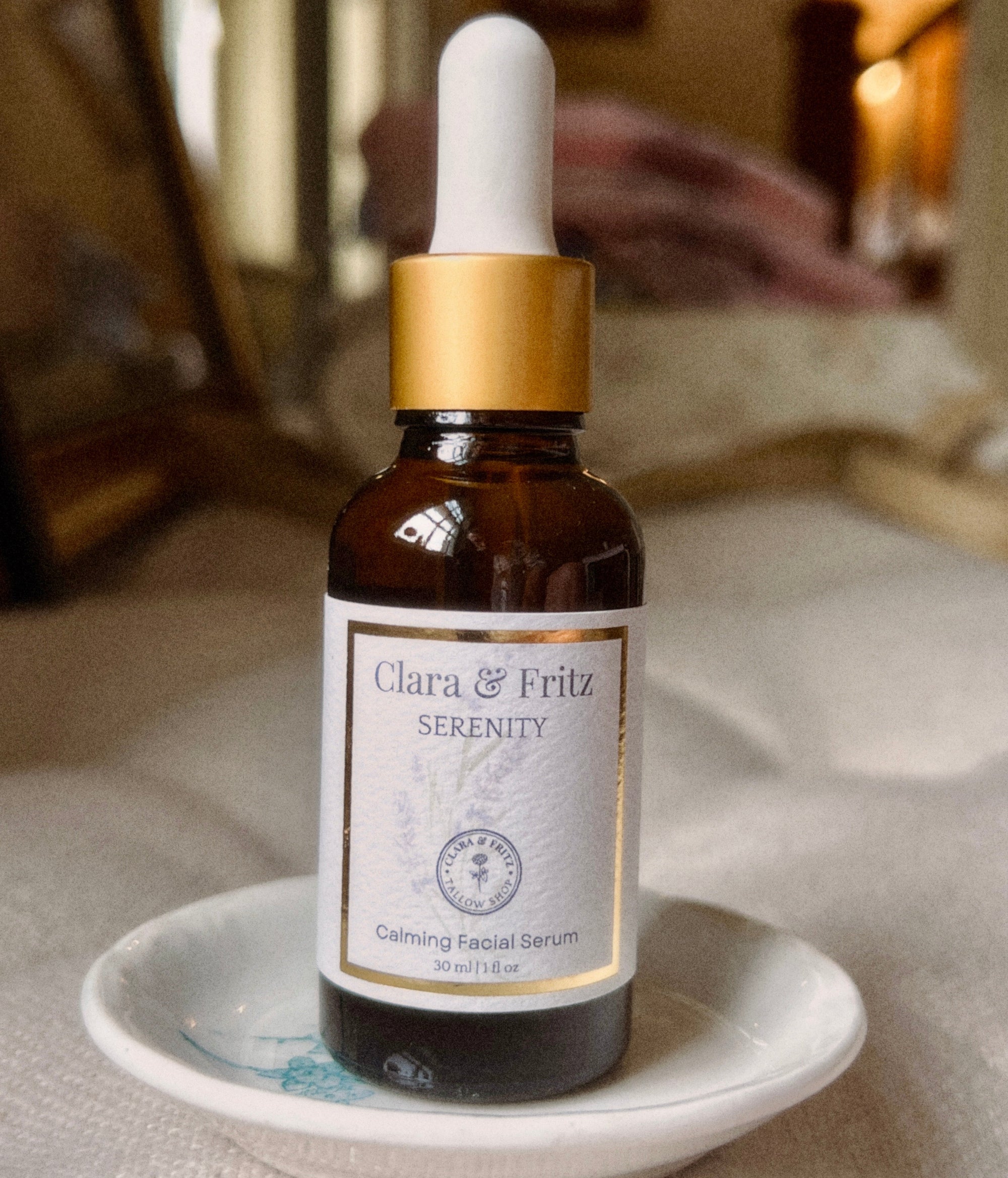A bottle of Clara &amp; Fritz Serenity Facial Serum in an amber glass dropper bottle with a white label and gold collar, resting on a small white dish against a softly blurred indoor background.