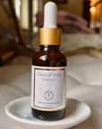 A bottle of Clara & Fritz Serenity Facial Serum in an amber glass dropper bottle with a white label and gold collar, resting on a small white dish against a softly blurred indoor background.